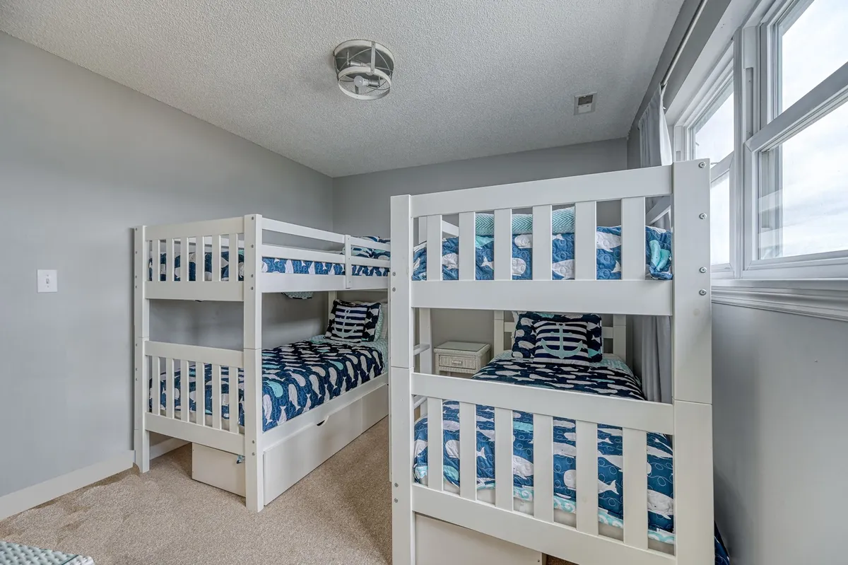 A cheerful bunkroom filled with natural light, featuring two twin-over-twin bunk beds with colorful bedding and playful decor. Large windows allow light to stream in, and the arrangement invites both rest and play for younger guests.