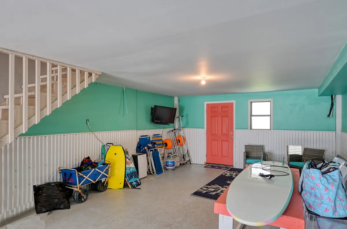 A playful and relaxed interior with a colorful door, a surfboard leaning against the wall, TV, and a stack of chairs. The vibe is casual and fun, reflecting a space where families can relax after beach adventures, catch up on TV, or prepare for the next outing.