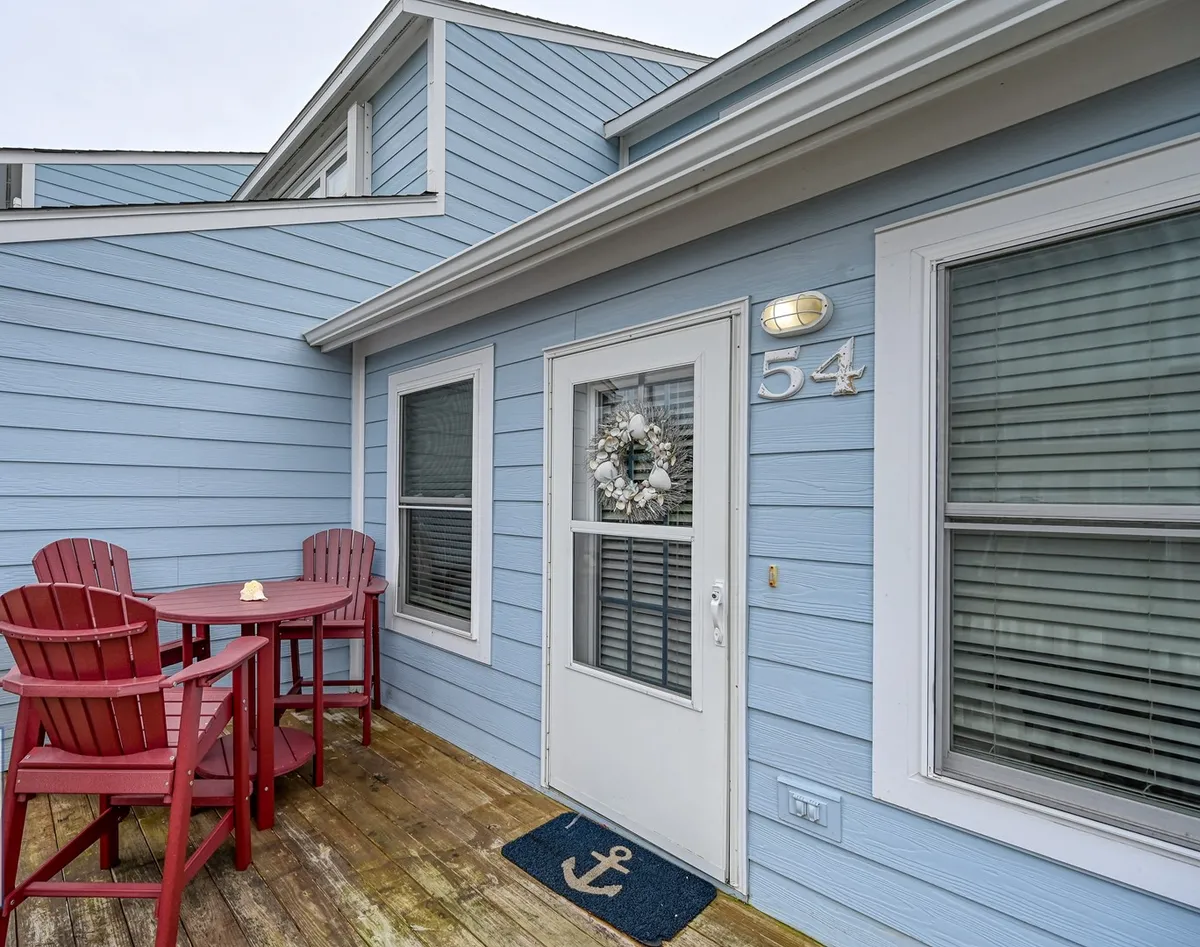 This exterior image showcases the blue exterior walls of Bermuda Landing, with red Adirondack chairs on the deck, a welcoming wreath adorning the door, and the house number 54 clearly visible. The inviting setup hints at a perfect spot for morning coffee, sunset drinks, or casual gatherings with friends as the sun sets over the sound.