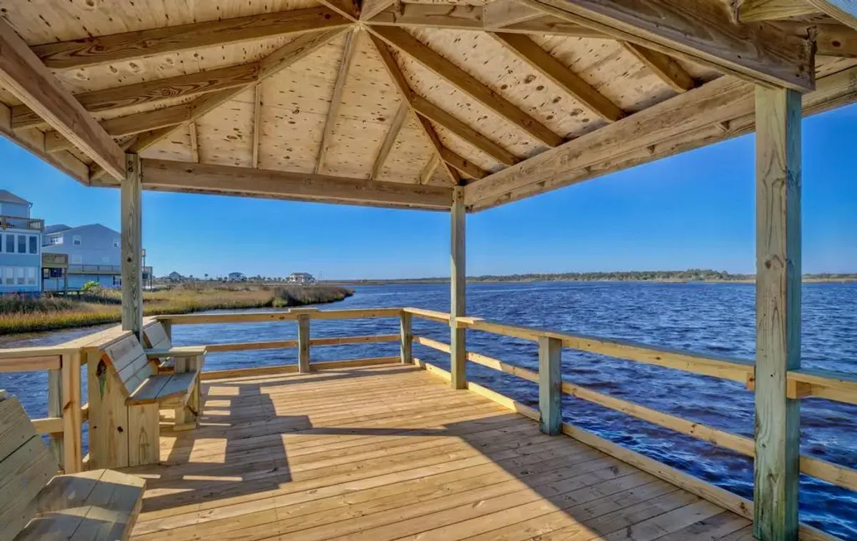 The image showcases a serene wooden deck overlooking tranquil waters, inviting seating perfect for relaxing or enjoying a morning coffee, and an expansive view ideal for watching sunsets. The setting exudes calmness and leisure, with the waterfront just steps away and hints of the surrounding natural beauty.