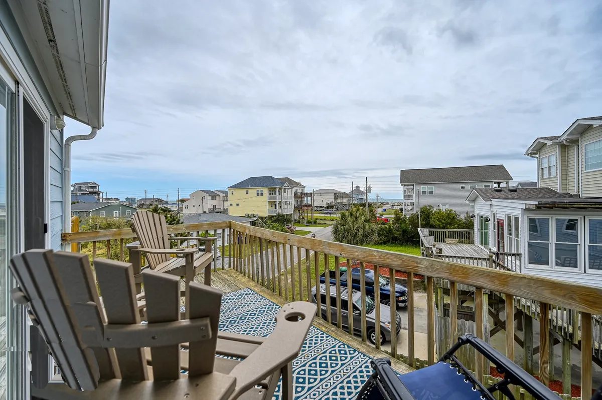 The image features a tranquil wooden deck with Adirondack chairs and a patterned rug, looking out toward neighboring houses. The perspective from the deck suggests a relaxing vantage point for watching sunsets or observing local wildlife, connecting the home to the outdoor environment and the soundfront.