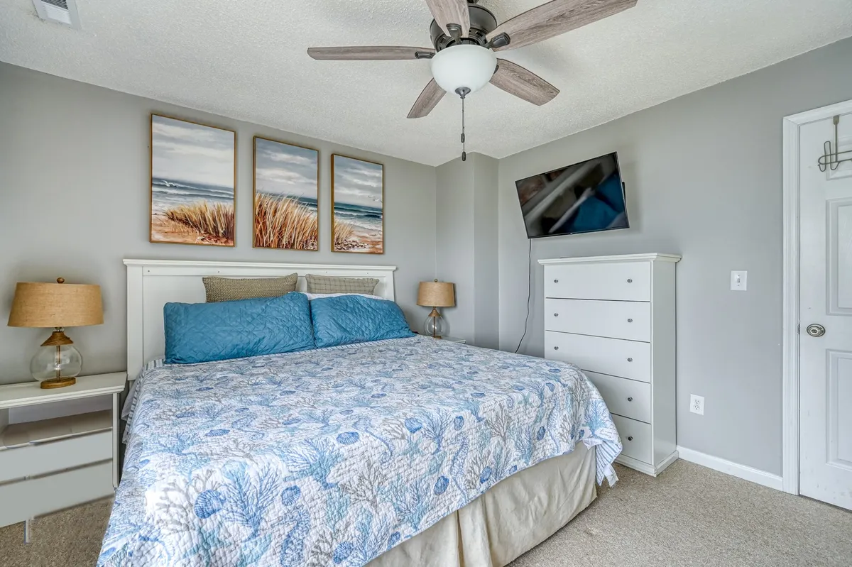 A tranquil king or queen bedroom featuring blue and white bedding, three beach-themed paintings above the bed, a nightstand with a lamp, a tall dresser, and a mounted TV. The space feels airy, with soft colors and natural light that create a peaceful environment for relaxation or unwinding at the end of the day.