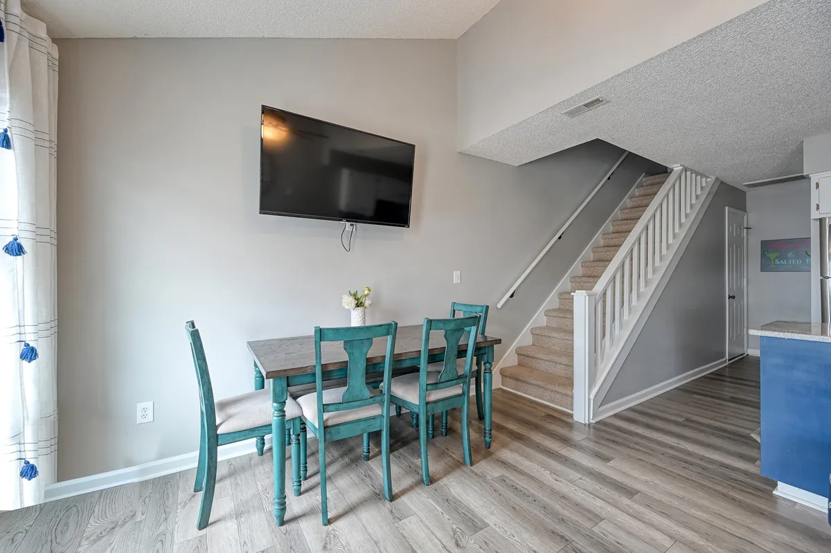 This image features an open-plan dining space with a teal-accented table and chairs, a wall-mounted television, and a staircase leading to the upper floor. A vase with flowers decorates the dining area, and the space is awash in natural light, highlighting its welcoming ambiance.
