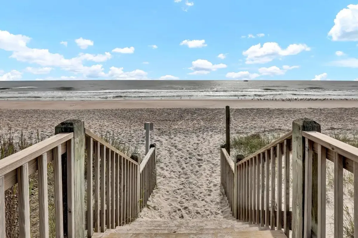 A stunning exterior view of wooden stairs leading directly to a broad, sandy beach with the expansive ocean in the distance and a beautiful blue sky overhead. The scene evokes the excitement of easy beach access and the tranquility of coastal vistas.