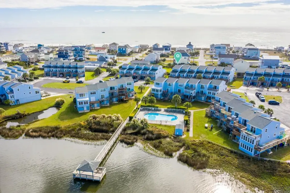 This exterior image captures a vibrant scene featuring a waterfront dock extending into the sound, a sparkling swimming pool in the foreground, and lush green lawns bordered by neighboring houses. There are people enjoying activities such as swimming, relaxing by the pool, and walking along the dock, all set against a softly glowing sunset.