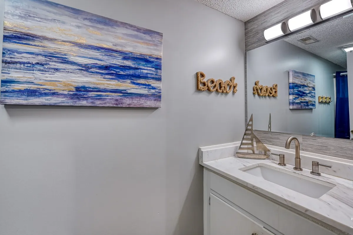 A serene bathroom scene highlighted by an abstract ocean painting and a wooden 'Beach' sign above a modern sink and faucet. The space is bright and airy, with details evoking a coastal sense of place and readiness for a day at the beach.