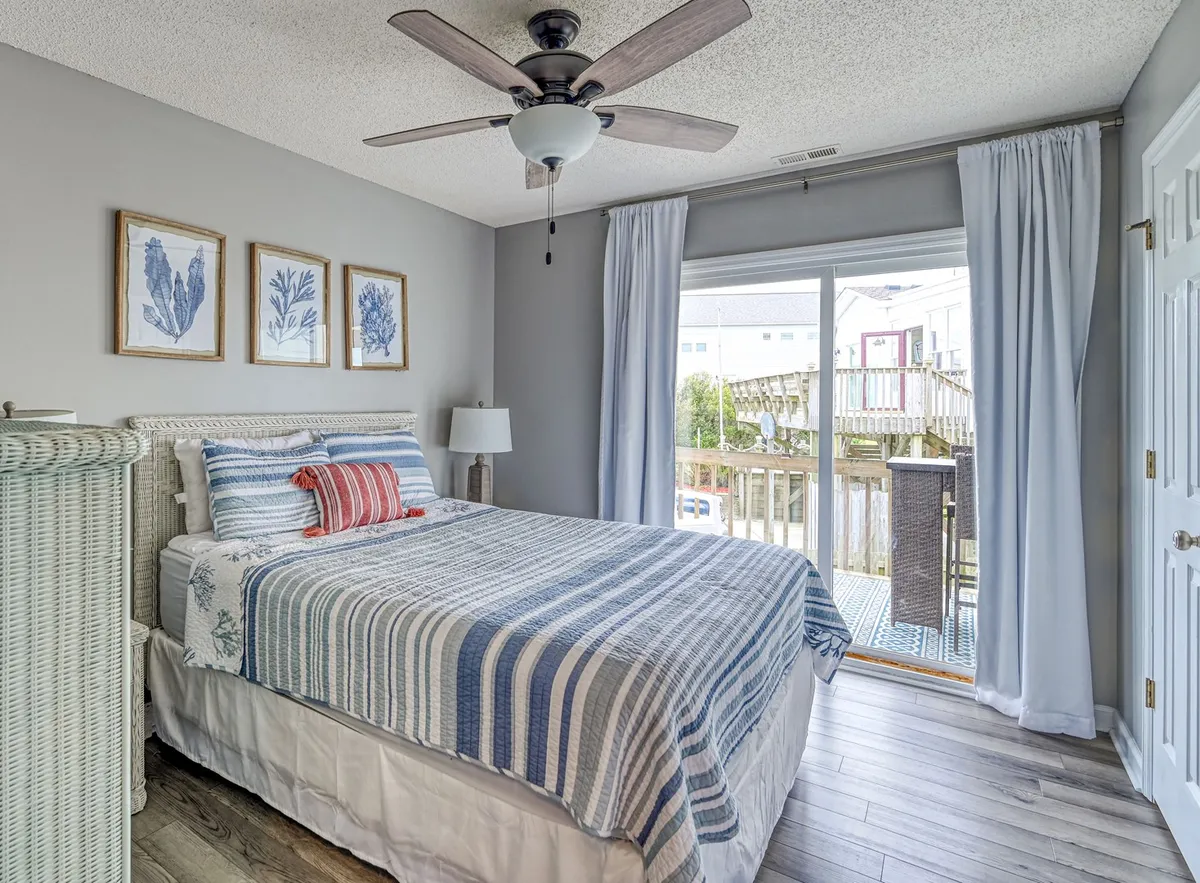 A queen or king bedroom with striped bedding, a sliding glass door leading to a deck or balcony, and framed wall art. Light fills the room, hinting at easy access to outdoor spaces and the enjoyment of fresh breezes and scenic views, in line with rooms that offer deck access.