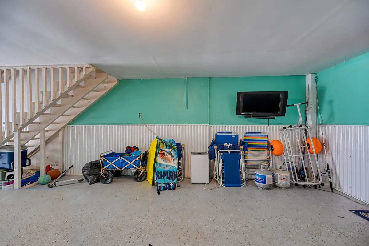 A practical ground-level space featuring a staircase, visible beach equipment, a television, coolers, and comfy seating. The area looks ready for families to store their beach gear, relax together after a day out, or catch a show on TV. The atmosphere is casual and functional, with storage solutions and entertainment options in one spot.